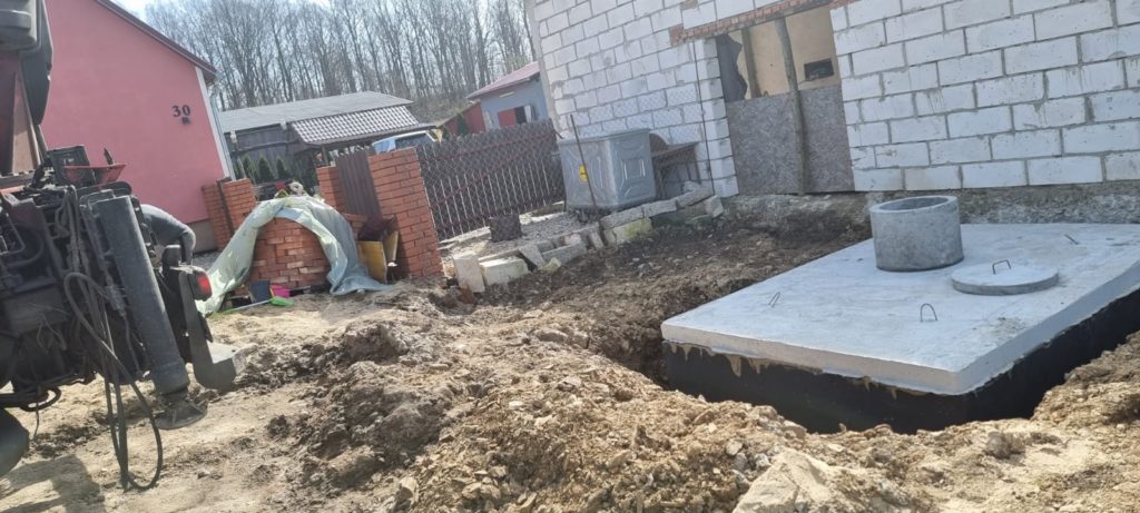 Wykop z częściowo zasypanym szambem betonowym obok murowanego budynku