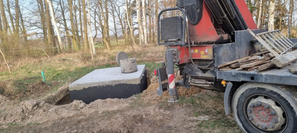 Szambo betonowe w wykopie, w tle samochód z dźwigiem i las